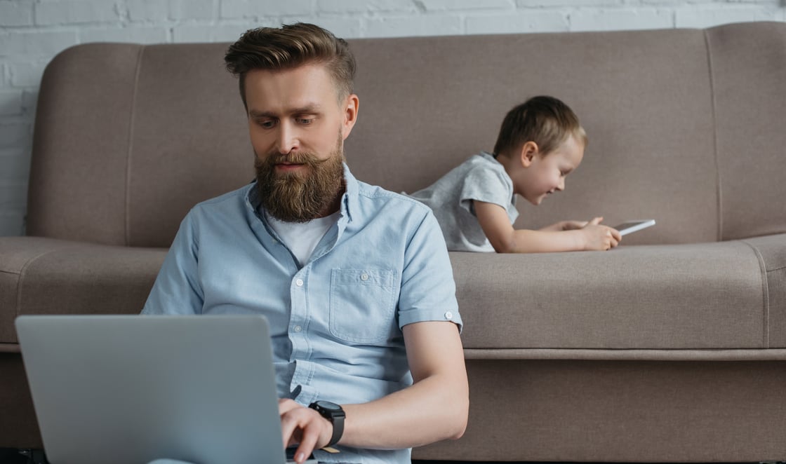 Man taking TREC Parenting Course on his laptop. Young son in background on his table.