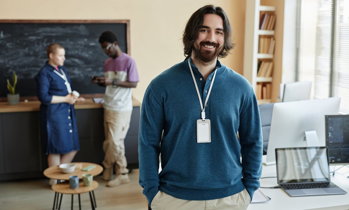 Young male teacher in classroom, smiling. Students in background.